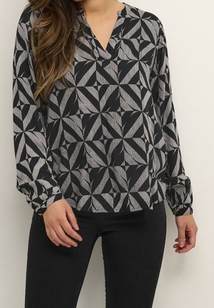 Frau trägt eine schwarz-grau gemusterte, langärmelige Bluse mit V-Ausschnitt, kombiniert mit schwarzen Hosen vor einem schlichten Hintergrund.