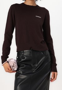 Person, die einen dunkelbraunen Pullover mit dem Schriftzug "2NDDAY" und einen hochgeschnittenen schwarzen Lederrock trägt, hält in einer Hand eine gefaltete hellrosa Tasche.