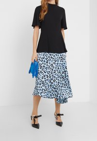 Czarny krótki top peplum z krótkim rękawem połączony z jasnoniebieską spódnicą midi w tygrysie plamki, trzymający niebieski frędzlowany dodatek i noszący czarne buty na wysokim obcasie.