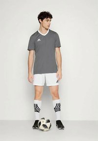 Szürke sportmez fehér gallérral, fehér sorttal és fekete focicipővel párosítva; csíkos fekete-fehér zokni, kezében egy focilabdával.