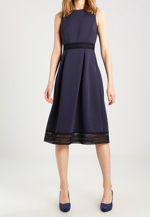 Robe midi bleu marine sans manches avec ceinture noire et ourlet en dentelle, portée avec des talons bleu marine assortis.