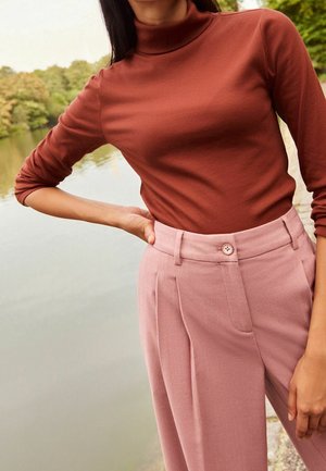 Vrouw in een rustkleurige coltrui en hoogopgesneden stofroze broek staande bij een kalm meer met bomen op de achtergrond.
