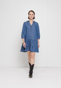 Robe en denim bleu avec un décolleté en V, des manches trois-quarts, des boutons au cou et un ourlet volanté. Portée avec des bottes de cowboy noires.