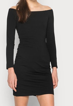 Kobieta w dopasowanej, czarnej sukience z długim rękawem i odsłoniętymi ramionami, stojąca na tle jednolitego, jasnego tła.