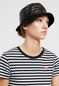 Pălărie neagră tip bucket cu o textură încrețită, având o margine complet neagră. Purtați cu un tricou cu dungi negre și albe, punând accent pe un stil lejer.