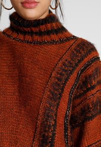 Suéter de punto en naranja oscuro con rayas negras; cuenta con un cuello alto acanalado y patrones texturizados a lo largo de las mangas.