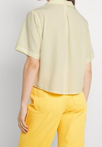 Chemise à manches courtes de couleur jaune pâle avec boutons, col plat, tissu lisse et ourlet court, associée à un pantalon jaune.