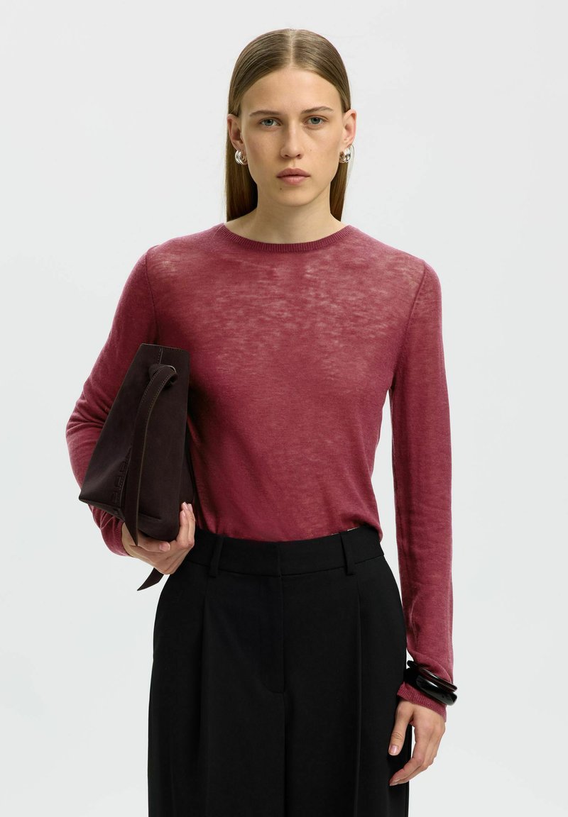 Femme aux cheveux raides portant un pull fin bordeaux et un pantalon noir, tenant un sac à main marron foncé dans un bras, devant un fond uni.