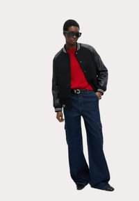 Chaqueta varsity negra con mangas de cuero, suéter de punto rojo y pantalones cargo de mezclilla anchos. Los accesorios incluyen gafas de sol negras y un cinturón.