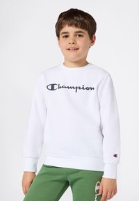 Gutt i hvit Champion hettegenser og grønne bukser, står mot en enkel hvit bakgrunn og smiler svakt.