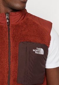 Chaleco de forro polar rojo con un bolsillo acolchado marrón. Presenta un alto cuello, cremallera completa y un logo bordado. Apariencia texturada y cálida.