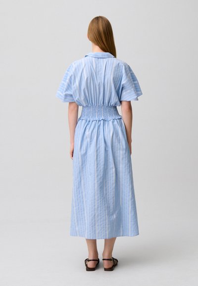 Donna con capelli lunghi e lisci che indossa un vestito midi a righe verticali blu e bianche con maniche larghe e sandali neri, in piedi davanti a uno sfondo neutro.