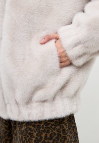 Giacca in pelliccia artificiale color beige chiaro con una texture morbida e una vita elastica. Presenta tasche laterali e polsini aderenti, abbinata a pantaloni con stampa leopardata marrone.