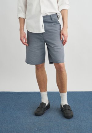 Άνδρας που φοράει γκρι σορτς, λευκό πουκάμισο με μακριά μανίκια, λευκές κάλτσες και μαύρα παπούτσια χωρίς κορδόνια, στέκεται πάνω σε μπλε χαλί.