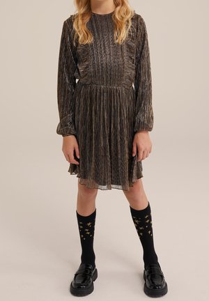 Dziewczyna w plisowanej, długorękawowej sukience o ciemnym metalicznym odcieniu, czarnych skarpetkach z motywem złotych gwiazd oraz czarnych błyszczących mokasynach stoi na neutralnym tle.