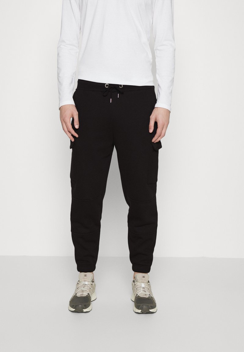 Zwarte sweatpants met een gladde textuur, voorzien van een trekkoordsluiting, zijzakken en een taps toelopende pasvorm. Gepaard met beige en olijfgroene sneakers.