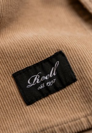 Brown corduroy fabric with a black label featuring white cursive text that reads "Reell" and "est. 1997." Textured, ribbed surface detail visible.