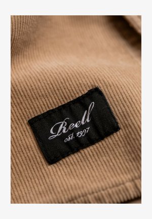Brown corduroy fabric with a black label featuring white cursive text that reads "Reell" and "est. 1997." Textured, ribbed surface detail visible.