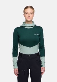 Junge Frau in einem grünen und hellblauen langärmeligen Outdoor-Oberteil und schwarzen Hosen, steht und schaut mit dem Gesicht nach vorne vor einem einfarbigen Hintergrund.