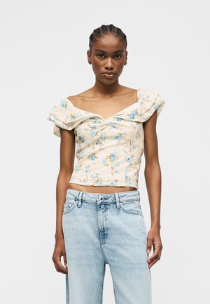 Γυναίκα με κρέμ φλοράλ μπλούζα χωρίς ώμους με βολάν και ανοιχτό μπλε τζιν ψηλόμεσο, στέκεται μπροστά σε απλό φόντο.