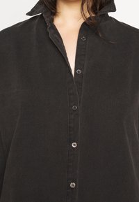 Camisa negra de botones con cuello doblado, que presenta una tela texturizada y cinco botones en la parte delantera. Diseño simple y clásico.