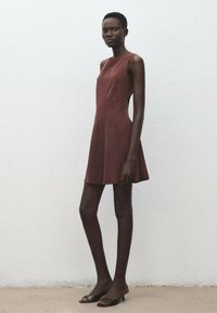 Vestido corto marrón sin mangas con un corsé ajustado y falda acampanada, con cortes laterales. La modelo lleva tacones negros destalonados.