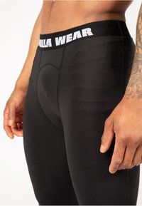 Zwarte sportleggings hebben een strakke pasvorm met een onderscheidende tailleband die in het wit is gemerkt met "Gorilla Wear". Ze hebben een gladde textuur en subtiele stiksels.