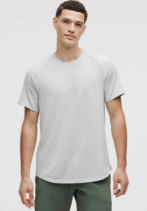 Mann in einem hellgrauen kurzärmligen Rundhals-Shirt und olivgrünen Hosen, der vor einem schlichten hellen Hintergrund steht.