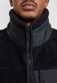 Czarna polarowa kurtka z wysokim kołnierzem, wyposażona w kontrastujący błyszczący materiał przy szyi, z pełnowymiarowym zamkiem błyskawicznym i fakturowaną powierzchnią.