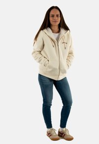Sudadera con cremallera en color beige, con un forro texturizado, dos bolsillos frontales y texto bordado. Combinada con jeans azules y zapatillas en tonos beige y marrón.