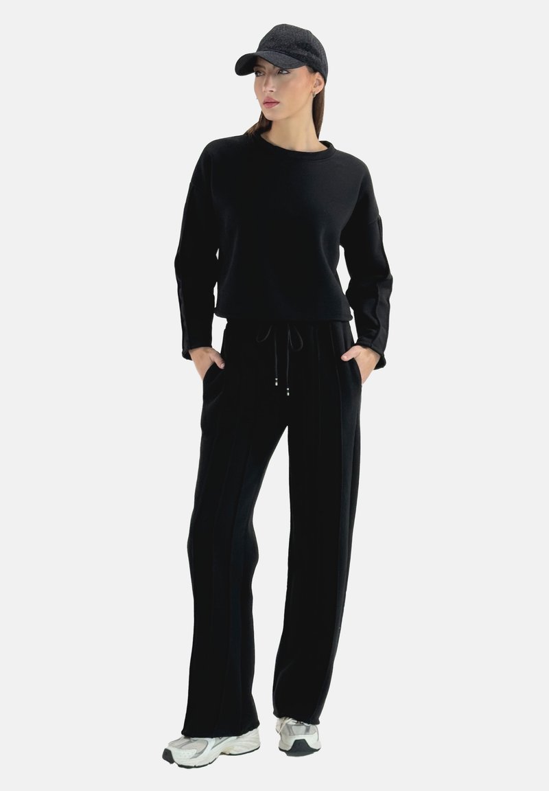 Felpa nera corta con maniche lunghe, abbinata a pantaloni neri con coulisse e righe verticali. Scarpe da ginnastica bianche e un cappellino nero.