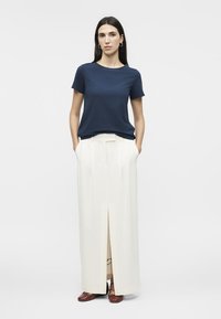 Γυναίκα στέκεται με τα χέρια στις τσέπες, φοράει μπλε ναυτικό κοντομάνικο πουκάμισο, μακριά λευκή φούστα με σκίσιμο μπροστά και καφέ παπούτσια, πάνω σε απλό λευκό φόντο.