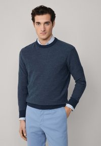 Mornarska pletena dukserica, okrugli izrez, rebrasti manšeti i donji rub, nosi se preko svijetloplavog prugastog košulje, u kombinaciji sa svijetloplavim hlačama.