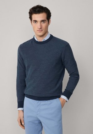 Marineblauer Strickpullover, runder Ausschnitt, gerippte Bündchen und Saum, über ein hellblau gestreiftes Hemd gezogen, kombiniert mit hellblauen Hosen.