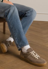 Baskets en daim marron avec des lacets crème, semelle en caoutchouc et un logo subtil. Portées avec un jean bleu clair et des chaussettes blanches sur un sol en bois.