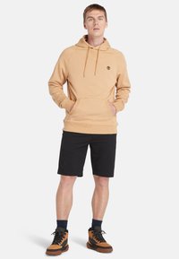 Beige hættetrøje med en frontlomme og snore, parret med sorte shorts og brune vandresko. Simple, afslappet design.