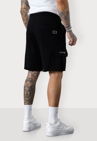Shorts cargo noirs avec un ourlet effiloché, dotés d'une poche latérale et d'un logo brodé blanc. Associés à des baskets blanches et des chaussettes côtelées.