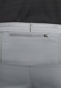 Sive hlače za tek z teksturiranim zaključkom, opremljene zatvorenim stražnjim džepom i bijelim elastičnim pojasom s oznakom "NIKE RUNNING."