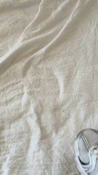 Tecido branco amarrotado com algumas manchas, mostrando uma textura suave. Um objeto de plástico transparente parcialmente visível no canto inferior direito.