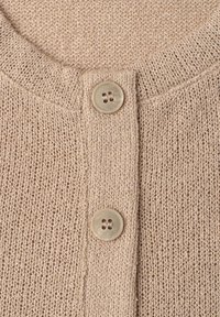 Cardigan beige en tricot avec un col rond et deux boutons beiges le long de la patte de devant.