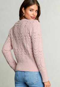 Maglione rosa a maglia con motivo in pizzo, polsini e orlo a coste, e maniche lunghe. Indossato con jeans in denim fitted blu chiaro.