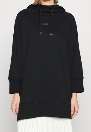 Personne portant un sweat à capuche noir avec cordons et petit logo blanc "Dondup" centré sur la poitrine, associé à une jupe plissée blanche.