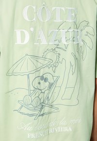Lichtgroene stof met een cartoon hond in een zonnebril die ontspant onder een parasol, met geborduurde tekst "CÔTE D'AZUR" en "FRENCH RIVIERA".