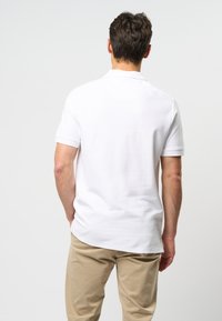 Polo shirt blanc à manches courtes avec une texture lisse et une coupe classique, associé à un pantalon beige clair, vu de l'arrière.