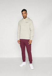 Světlý béžový mikina s přední kapsou a stahovacími šňůrkami, doplněná vínově červenými zužujícími se tepláky a bílými teniskami. Na mikině je viditelné logo.