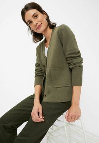 Olivengrüner Cardigan mit V-Ausschnitt, gerippten Bündchen und einer einzelnen Fronttasche. Das Material scheint strukturiert zu sein. Wird über einem weißen Hemd getragen.