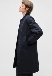 Tmavomodrý dlhý kabát s prešívanou podšívkou, bočnými vreckami a vreckom na ramene so zipsom. Má zapínanie na gombíky vpredu a hladký, matný povrch.