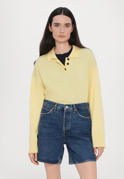 Maglione polo giallo a maglia con colletto e tre bottoni, abbinato a pantaloni shorts in denim scuro a vita alta. Design semplice e casual.