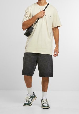 Beige katoenen T-shirt met grafische details, gecombineerd met donkere denim shorts. Een zwarte crossbody tas en groene Nike sneakers maken de look compleet.