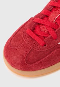 Röd mockasneaker med texturerad ovandel och rund tå. Har röda snören och en gummisula med en lätt genomskinlig kant.